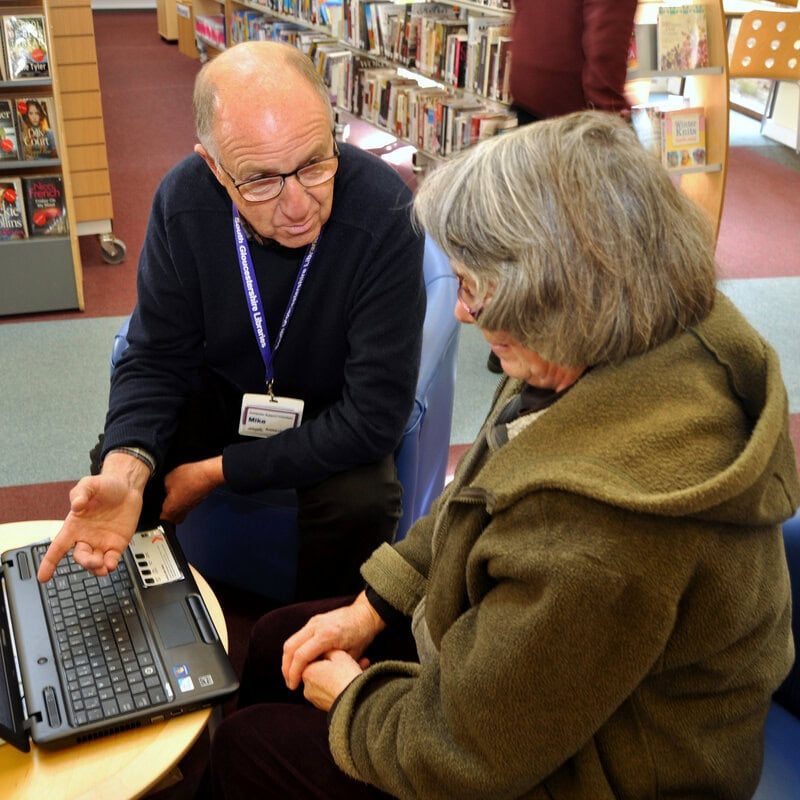 Local library volunteer A local library volunteer helps a woman with an IT question.